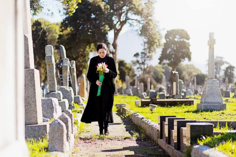 A bereaved young woman, wearing black and carrying flowers, walks through memorial stones in a cemetery.